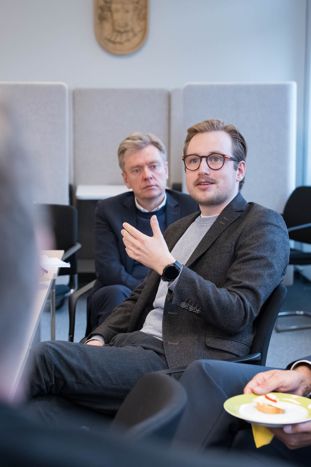 Ein Mann mit Brille gestikuliert. Im Hintergrund schaut ihm ein weitere Mann dabei zu.